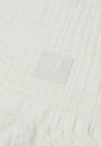 Witte geweven deken met een gestructureerd patroon, voorzien van franjes en een vierkante, lichtgrijze label met de tekst "Jollein".