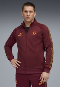 Mørkerød atletisk zip-up jakke med sorte detaljer, orange logoer og mønstrede sidestriber. Har en glat tekstur og en tætsiddende pasform.