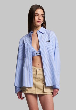 Blue and white striped oversized shirt, button-up design, two front pockets, worn over a matching blue crop top and beige mini skirt.