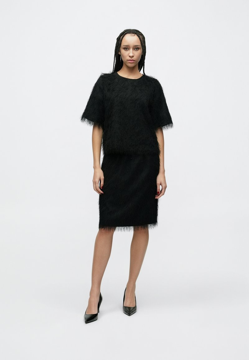 Tenue noire texturée comprenant un haut à manches courtes et une jupe mi-longue. Le tissu a un aspect duveteux, avec des talons noirs pointus.