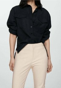 Chemise noire à boutons avec deux poches poitrine, manches retroussées et coupe décontractée, assortie à un pantalon tailleur beige clair.