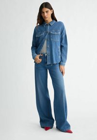 Veste en denim bleu clair à manches longues et devant boutonné ; associée à un jean large. Haut gris en dessous. Chaussures rouges à talons pointus.