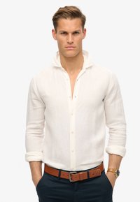 Camisa de lino blanca de botones con cuello doblado y mangas remangadas, combinada con un cinturón de cuero marrón y pantalones azul marino.