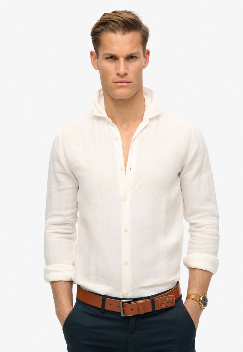 Camisa de lino blanca de botones con cuello doblado y mangas remangadas, combinada con un cinturón de cuero marrón y pantalones azul marino.