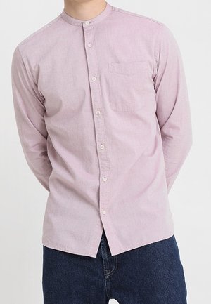 Mann trägt ein hellrosa langärmliges Hemd mit Mandarinkragen und dunklen blauen Jeans vor einem einfarbigen Hintergrund.