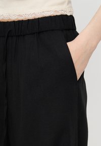 Χέρι που ακουμπά στην τσέπη μαύρου παντελονιού με ελαστική μέση, φορεμένο με κρεμ τοπ με δαντέλα σε ουδέτερο φόντο.