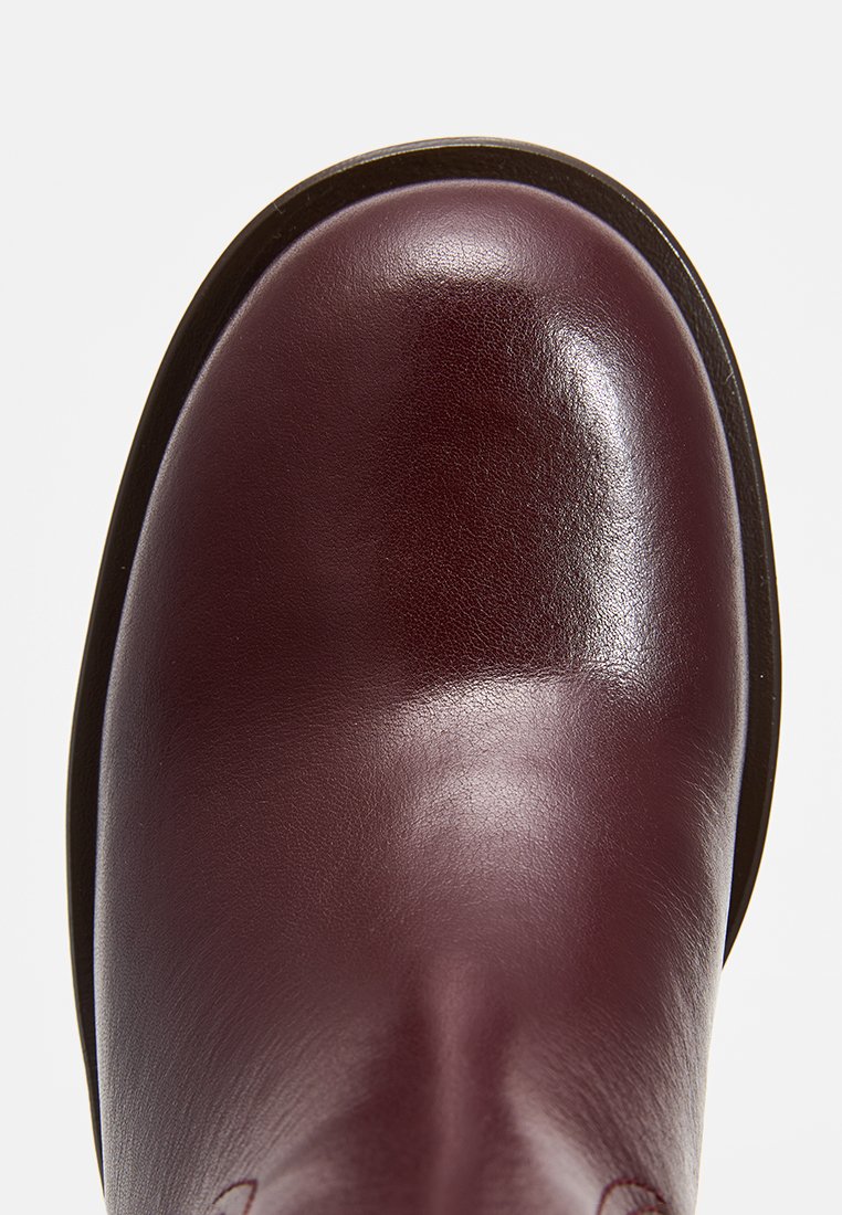 Botín de piel burdeos con una superficie lisa, punta redondeada y una suela resistente, que presenta costuras sutiles y un acabado brillante.