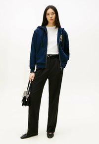 Marineblå zip-up hoodie med kænguruposer over en hvid T-shirt, parret med sorte vide bukser og sorte flade sko, mens der holdes en sort taske.