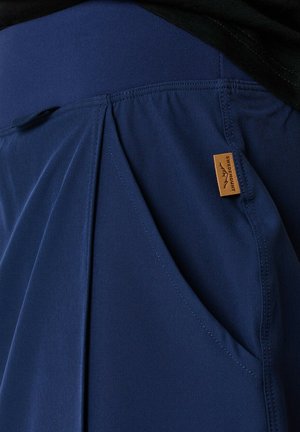 Pantaloni leggeri blu navy con una texture liscia, caratterizzati da un design plissettato e tasche laterali, etichettati con un piccolo cartellino beige.