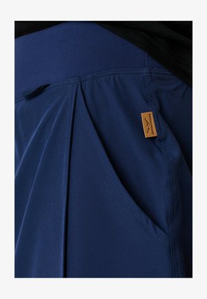 Pantaloni leggeri blu navy con una texture liscia, caratterizzati da un design plissettato e tasche laterali, etichettati con un piccolo cartellino beige.