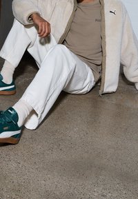 Buty sportowe Puma w kolorze zielononiebieskim z białymi akcentami, beżowa teksturowana kurtka, jasnobeżowa koszula i białe spodnie; siedzący na szarej betonowej podłodze.