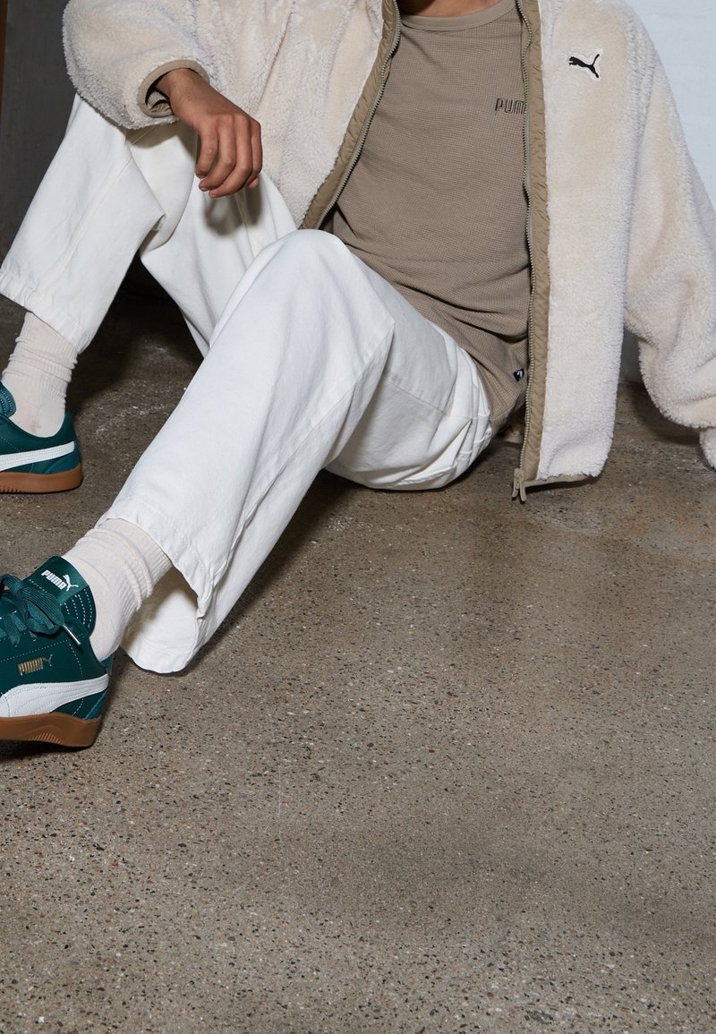 Buty sportowe Puma w kolorze zielononiebieskim z białymi akcentami, beżowa teksturowana kurtka, jasnobeżowa koszula i białe spodnie; siedzący na szarej betonowej podłodze.