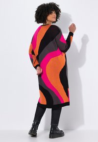 Femme aux cheveux bouclés portant une longue robe pull multicolore avec des motifs rose, orange, noir et gris, un pantalon noir et des bottes noires.