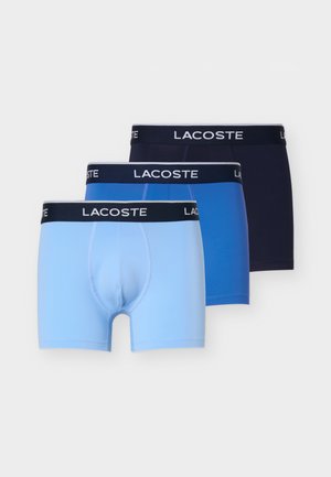 Kolm paari meeste bokserpükse helerohelises, keskmise sinises ja mereväesinis, igaüks musta vöökohaga, millel on Lacoste'i logo.