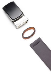 Metaliczna srebrna klamra z czarną tarczą, towarzysząca brązowo-czarnemu skórzanemu bransoletowi oraz długiemu czarnemu skórzanemu paskowi z nacięciami.