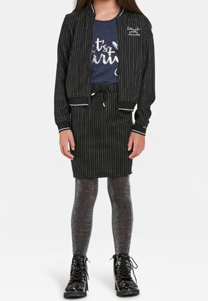 Fille portant une veste et une jupe rayées noires, un t-shirt graphique bleu marine, des collants gris scintillants et des bottines noires à lacets.