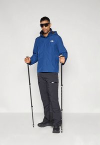 Homem com uma jaqueta azul e calças pretas segurando bastões de caminhada, usando óculos de sol pretos, em pé contra um fundo claro e liso.