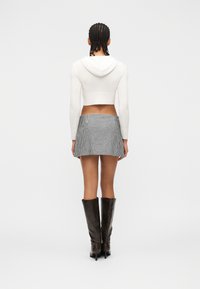 Personne aux cheveux tressés portant un sweat à capuche court blanc, une mini-jupe à carreaux noir et blanc, et des bottes noires jusqu'au genou, debout de dos.