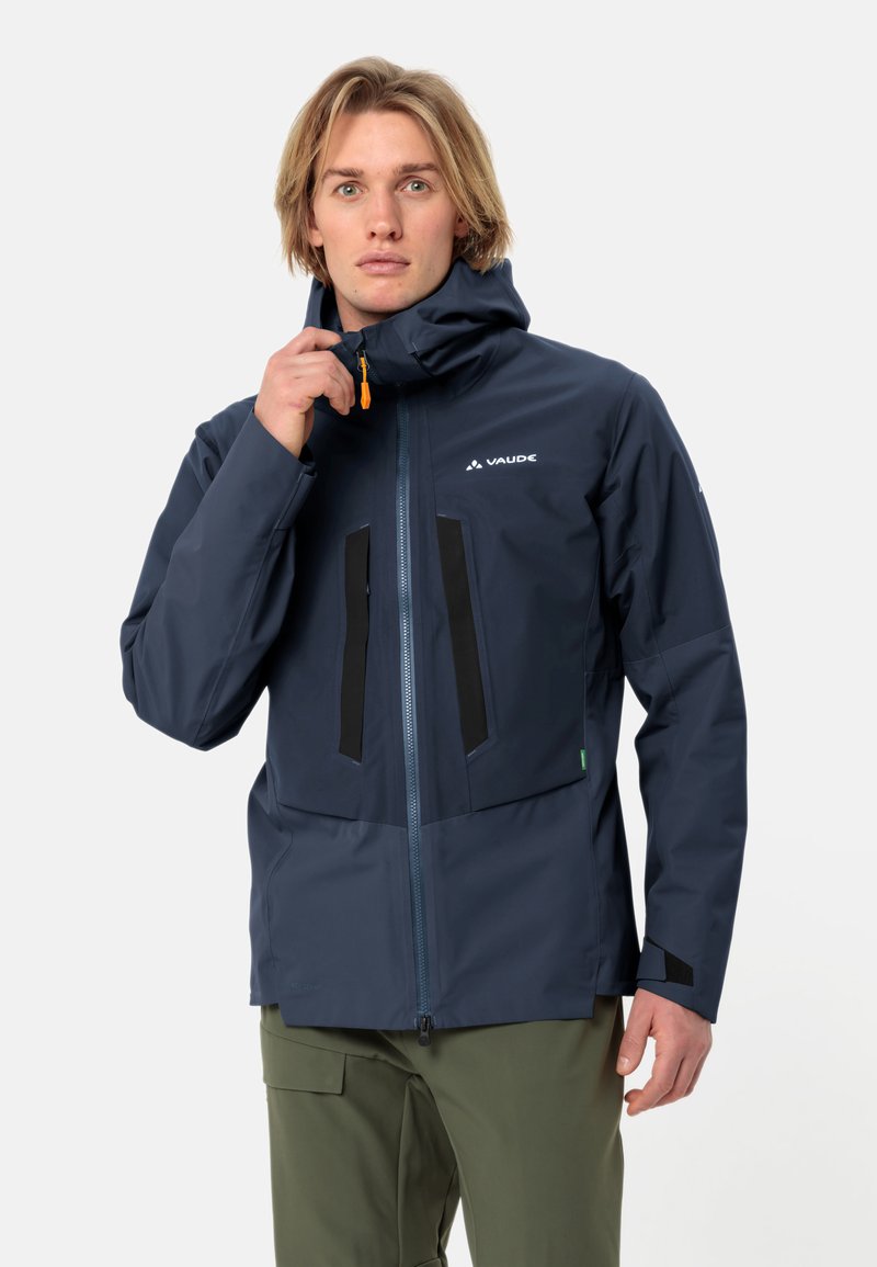 Giacca impermeabile blu navy con cappuccio, con accenti neri e tasche con zip. Realizzata in un materiale texturizzato e resistente, progettato per l'uso all'aperto.