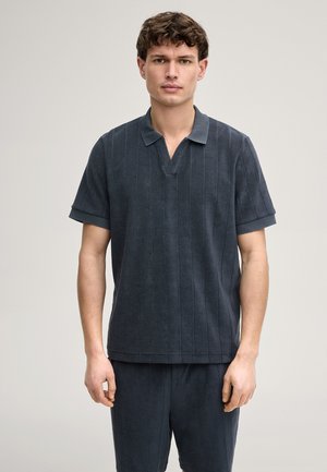 Jonge man met kort krullend haar, gekleed in een donkergrijs poloshirt met korte mouwen en bijpassende broek, tegen een effen lichte achtergrond.