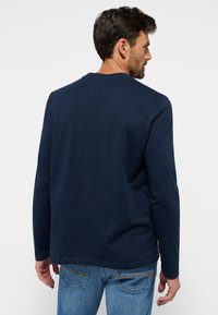 Tengerészkék, hosszú ujjú pamut póló, kerek nyakkivágással és egyenes aljjal. Kék farmernadrággal viselve, diszkrét varrási részletekkel.