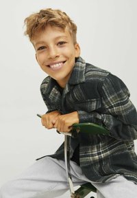 Rutig ullskjorta i mörkblått med vita och grå mönster, bärs över en svart topp, tillsammans med ljusgrå byxor. Sitter på ett grönt föremål.