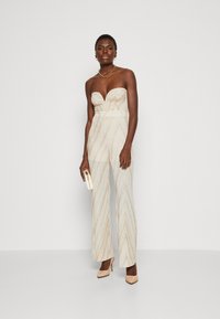 Beiger schulterfreier Jumpsuit mit einem strukturierten Zickzackmuster und metallischen Akzenten. Ausgestellte Beine und kombiniert mit einer goldenen Halskette und einer Clutch.