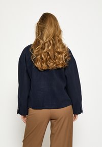 Külmalt meresinisest lahtise lõikega jope, millel on lame krae ja pikad varrukad, koos pruunide kohandatud pükstega.