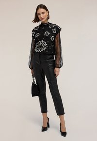 Blusa negra bordada con patrón floral y mangas de encaje transparente, combinada con pantalones de cuero sintético negros ajustados y tacones altos negros.