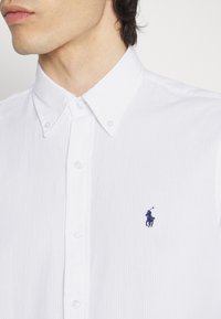 Vit skjorta med knappar som har en texturerad tyg, klassisk krage och en liten navy polo-logotyp p å vänster sida.