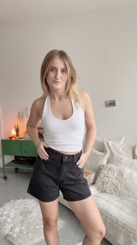 Crne traperice šorts s visokim strukom i prednjim kopčanjem na gumb, kombinirane s uskim bijelim halter topom, prikazane u svijetloj prostoriji.