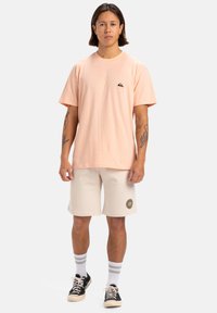 Camiseta rosa claro de manga corta con un parche de logo, combinada con pantalones cortos crema que tienen un logo, calcetines blancos con rayas grises y zapatillas negras.