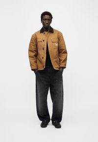 Άνδρας που φορά καφέ μπουφάν με μαύρο κολάρο, μαύρο πουκάμισο, φαρδιά ξεθωριασμένα μαύρα παντελόνια και μαύρα παπούτσια, στέκεται μπροστά σε απλό λευκό φόντο.