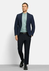 Marineblazer über einem hellgrünen Hemd mit Knopfleiste, kombiniert mit schwarzen Hosen und Schuhen. Glatter Stoff, taillierte Passform und elegantes Design.