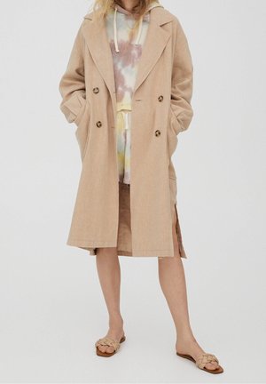 Femme portant un trench coat beige sur un sweat à capuche tie-dye pastel et un short, avec des sandales plates beige tressées, debout devant un fond uni.
