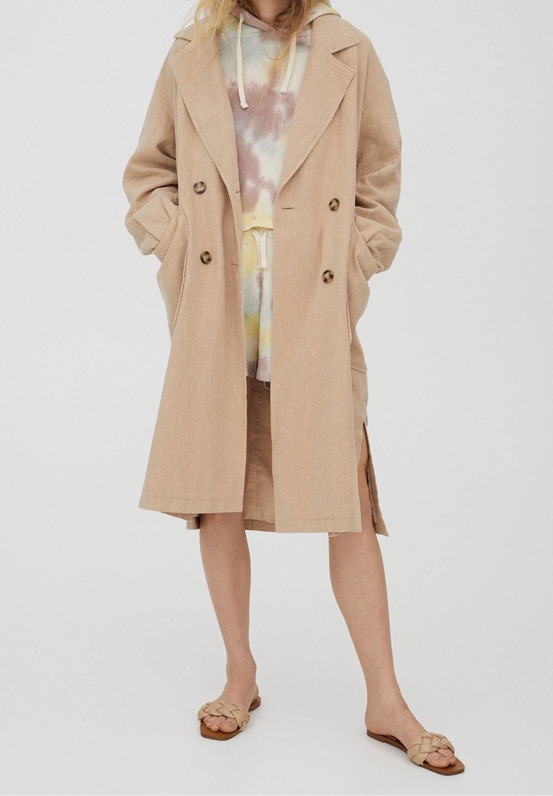 Femme portant un trench coat beige sur un sweat à capuche tie-dye pastel et un short, avec des sandales plates beige tressées, debout devant un fond uni.