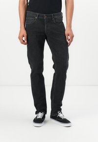 Jeans de mezclilla negra con un ajuste slim, diseño de cinco bolsillos y detalles en botón marrón, combinados con zapatillas negras y cordones blancos.
