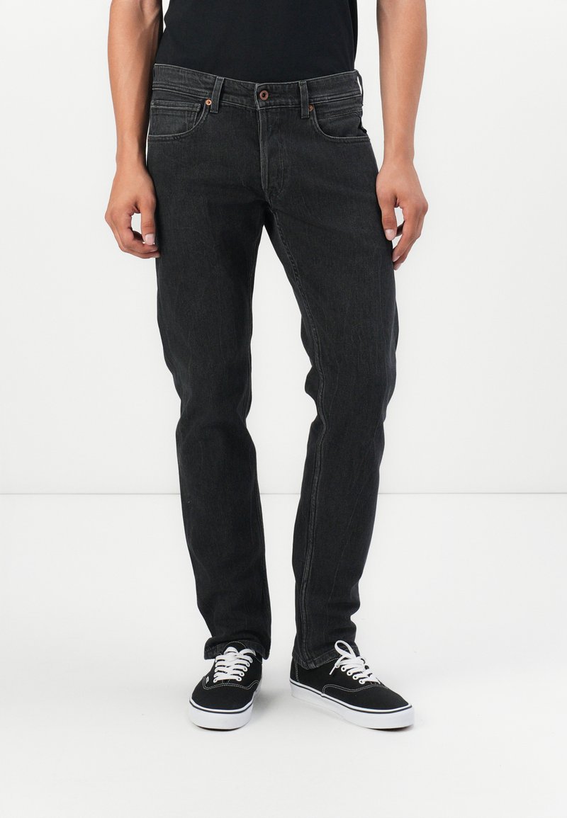 Jeans de mezclilla negra con un ajuste slim, diseño de cinco bolsillos y detalles en botón marrón, combinados con zapatillas negras y cordones blancos.