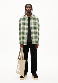 Grün kariertes Hemd mit Knopfverschluss, schwarzes T-Shirt, schwarze Cordhosen und eine beige Tragetasche mit einem weißen Logo. Schwarze Sneakers.