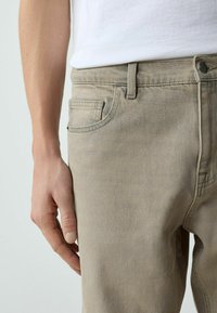 Rechte Hand ruht neben beigen Denimhosen, die mit einem weißen, hineingesteckten Hemd getragen werden, wobei Vorderhosentasche und Knopfdetails sichtbar sind.