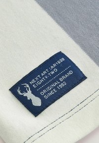 Grijs en crèmekleurig gestreept stof met een blauw label met witte tekst en een hertgrafiek. Zachte textuur en nette stiksels aan de randen.