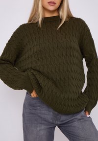 Grüner gestrickter Pullover mit strukturiertem Muster, mit einem gerippten Halsausschnitt und langen Ärmeln, getragen über grauen Jeans.