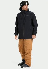 Μαύρο μπουφάν σκι με ψηλό γιακά και φερμουάρ, σε συνδυασμό με καφέ cargo παντελόνι με τσέπες. Τα μποτάκια είναι μαύρα με κομψό σχέδιο.