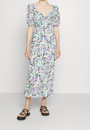 Vrouw draagt een midi-jurk met paarse, blauwe en witte bloemen, pofmouwen en zwarte platte sandalen met bandjes en staat tegen een effen achtergrond.