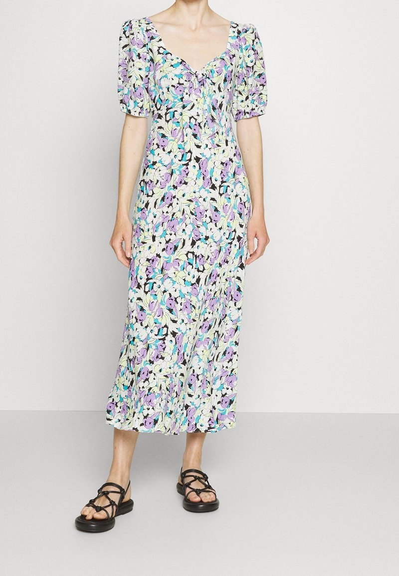 Femme portant une robe midi fleurie violette, bleue et blanche avec des manches bouffantes et des sandales plates noires à brides, debout devant un fond uni.