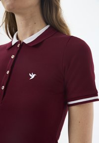 Burgundské polo tričko s bílým límečkem a akcenty, s malým bílým logem ptáka. Materiál má texturovaný povrch a zapínání na knoflíky.