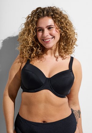 Usmievavá žena s kučeravými blond vlasmi, oblečená v čiernej podprsenke a zodpovedajúcich čiernych nohavičkách, stojaca pred bielym pozadím.