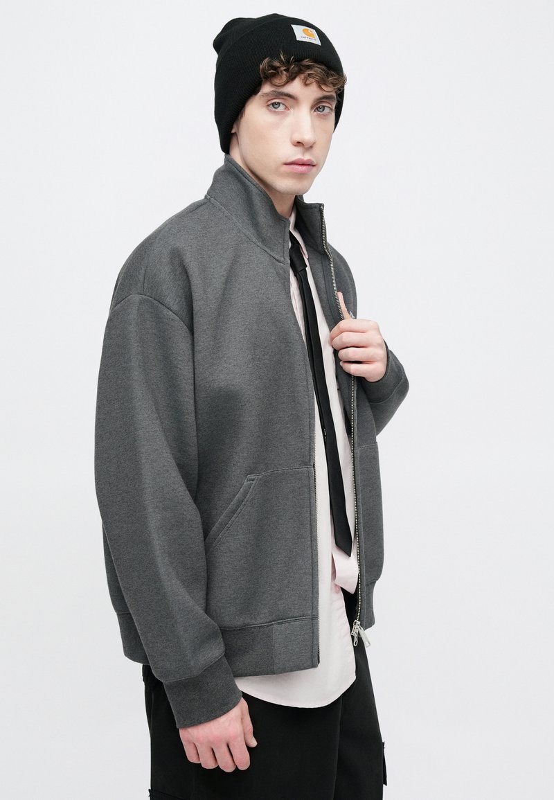 Veste en polaire grise avec un col montant, une fermeture éclair à l'avant et des poches latérales, portée avec un bonnet noir et une chemise à col en dessous.