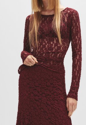 Femme portant un haut en dentelle bordeaux à manches longues et une jupe assortie à motifs floraux, tenant l'ourlet du haut légèrement relevé.