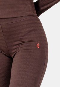 Close-up van bruine, getextureerde leggings met een klein rood geborduurd symbool met een hart op de bovenste dij.